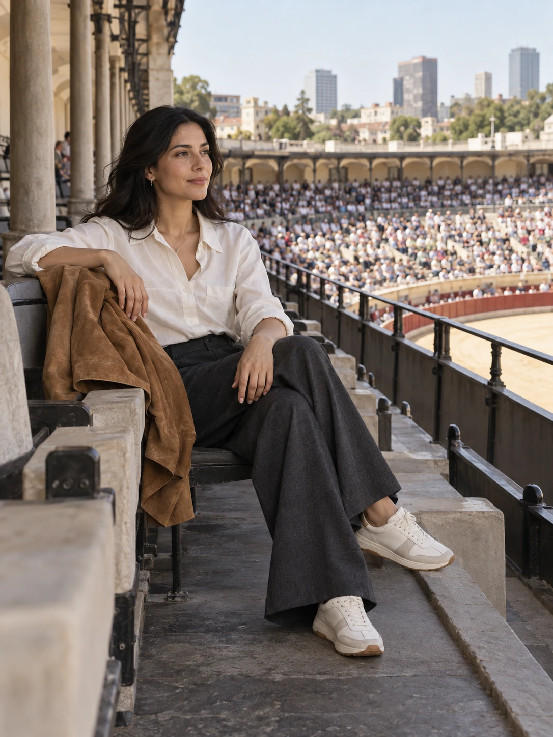 A woman in an ivory cotton shirt, wide charcoal trousers and supportive trainers seated in shaded stadium seating, with a tobacco jacket over her arm.
